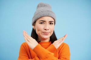 Disappointed korean woman shows cross sign, stop gesture, disapprove and rejects smth, refusing, standing over blue background photo