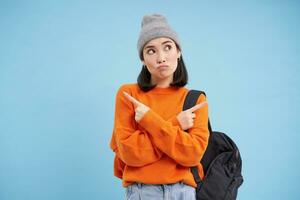 Puzzled urban girl, asian woman points sideways and thinking, making decision, choosing between two offers, standing over blue background photo