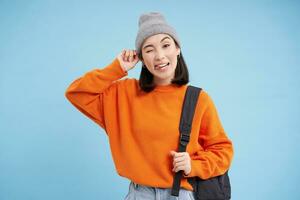 Beautiful modern korean woman in warm hat, holds bag, winks and smiles at camera, blue studio background photo