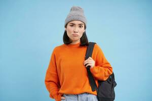 Young asian woman in hat, holds backpack, sulks and looks confused, perplexed emotion, stands over blue background photo