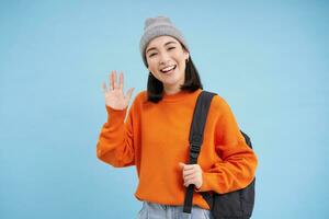 Friendly girl says hello, holds backpack and waves hand, greets you and laughs, stands over blue background photo