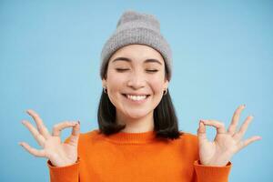 Sounds mind in healthy body. Smiling calm and relaxed asian girl in beanie, shows zen, relaxation gesture, meditating, standing over blue background photo