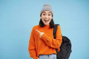 Enthusiastic asian woman, points finger left at banner, shows promo offer, smiles and looks happy, stands in hat with backpack, blue background photo