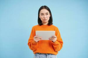 Woman with digital tablet thinking, looking aside with thoughtful face expression, standing over blue background photo