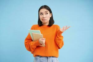 Confused asian woman, student with puzzled face shrugs shoulders, holds notebook, stands over blue background photo