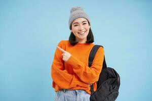 Stylish korean woman in hat, holds backpack, points finger at advertisement, shows promo offer banner, stands over blue background photo