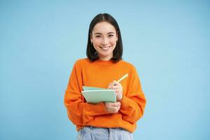Smiling cute girl student, making notes, writing down homework, studying, standing over blue studio background photo