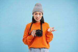 Portrait of asian woman in hat, holding digital camera with confused face, unprofessional photographer doesnt know how to take pictures on digicam, blue background photo