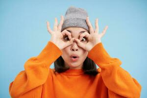 Close up portrait of funny Chinese girl, looks through hand glasses with surprised face expression, standing over blue background photo
