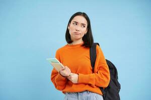 Bored college student, girl with backpack and notebooks, sulks and rolls eyes, stands upset against blue background photo