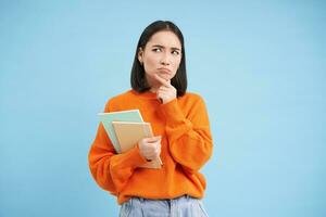 Thinking girl, student with notebooks, stands over blue background thoughtful, making decision photo