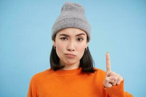 Asian girl in hat scolding you, shakes finger in disapproval, disappointed by behavious, standing over blue background photo