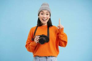 Surprised asian girl in hat, holds digital camera, looks amazed and impressed, poses over blue studio background with space for banner photo