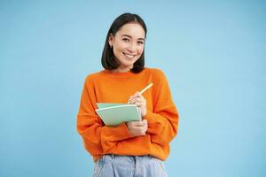 Education and students. Happy asian woman, holding notebooks and laughing, smiling at camera, enjoys going to University or College, blue background photo