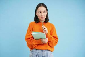 Thoughtful asian woman, student smiling, holding pen and notebook, thinking, inspired to write something, standing over blue background photo