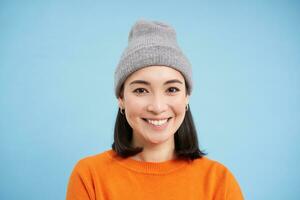 Close up portrait of smiling asian woman in warm hat, looking happy and cute at camera, has clear natural skin, stands over blue background photo