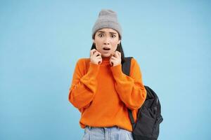 Portrait of korean woman looks scared, jumps terrified, stands with backpack in street clothing, blue background photo