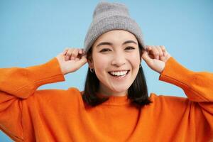 Close up portrait of happy, stylish korean girl puts on warm hat, smiles and looks joyful, stands against blue studio background photo