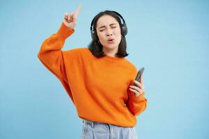 hermosa coreano mujer en auriculares, baile, escuchando música en móvil teléfono aplicación, sonriente, posando terminado azul antecedentes foto