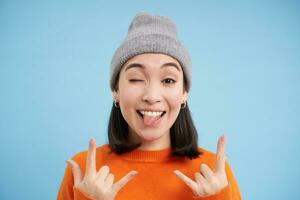 Close up portrait of funny and carefree asian girl in beanie, shows rock n roll, heavy metal gesture and tongue, stands over blue background photo