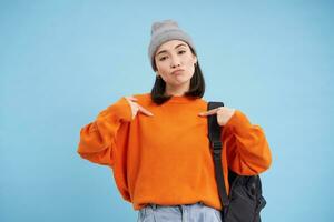 Stylish korean woman points at herself, stands in warm hat and holds backpak on shoulder, blue background photo