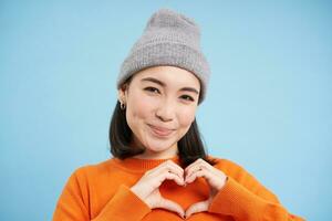 Close up of lovely asian girl in beanie, shows heart sign, looks cute, stands over blue studio background photo