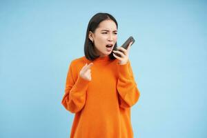 enojado asiático mujer grita a móvil teléfono, gritos en teléfono inteligente con furioso rostro, soportes terminado azul antecedentes foto