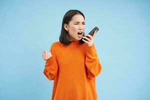 Angry asian woman yells at mobile phone, shouts in smartphone with furious face, stands over blue background photo