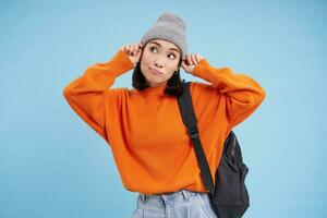 Happy stylish korean woman puts on hat, goes for walk with backpack, stands over blue studio background photo