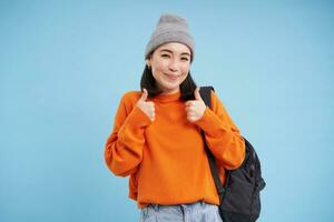 Cute korean woman shows thumbs up, approves smth good, likes and gives good feedback, recommends something, standing over blue background photo