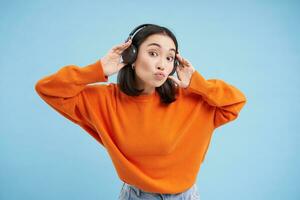 Beautiful korean woman in headphones, dancing and listening music in earphones, standing over blue background photo