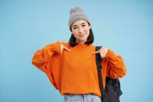 Stylish korean woman points at herself, stands in warm hat and holds backpak on shoulder, blue background photo