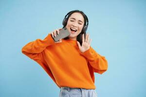 hermosa coreano niña canto en teléfono inteligente micrófono, escucha música en auriculares, canta karaoke en móvil aplicación, soportes terminado azul antecedentes foto