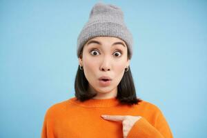 Close up portrait of korean girl in winter hat, points at herself, stands in orange sweatshirt over blue background photo