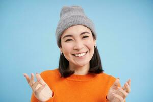 Close up portrait of happy smiling asian woman shows open hands, pointing, looking friendly, standing over blue background photo