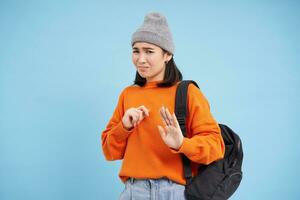 Asian girl grimaces, shows block hands gesture, stop sign, asks to stay back, rejects smth disgusting, stands over blue background photo