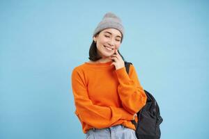 Modern college girl in hat, holds backpack on shoulder, smiles and laughs, stands over studio background photo