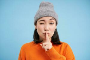 Shh its secret. Close up portrait of asian woman in hat, puts finger on lips, asks to keep quiet, hushing, standing over blue background photo