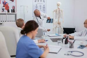 Physician specialist doctor explaining body anatomy structure using radiography and skeleton during bone examination in conference meeting room. Hospital group working at diagnosis treatment photo
