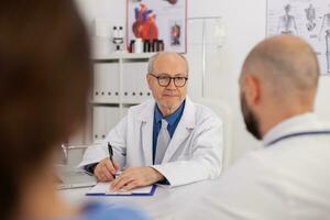 retrato de médico mayor hombre que se discute con hospital trabajo en equipo trabajando a médico pericia que se discute pastillas tratamiento. médico sentado a escritorio en conferencia reunión habitación diagnosticando enfermedad foto