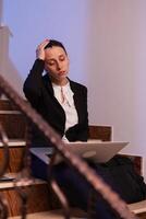 Stressed overworked businesswoman doing overtime for project deadline at late hours. Serious entrepreneur working on job project sitting on staircase of business building at night hours for job. photo
