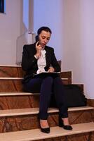 Overworked businessoman disccusing about project deadling on smartphone late at night doring overtime. Serious entrepreneur working on job project sitting on staircase of business building at night hours for job. photo