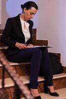 Overworked stressed businesswoman doing overtime for deadline project. Serious entrepreneur working on job project sitting on staircase of business building at night hours for job. photo