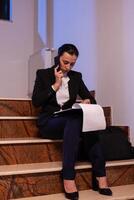 Stressed tired businesswoman readling project deadline during conversation on smartphone. Serious entrepreneur working on job project sitting on staircase of business building at night hours for job. photo
