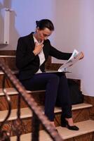 Overworked tired businesswoman reading financial raport of deadline project in the evening. Serious entrepreneur working on job project sitting on staircase of business building at night hours for job. photo