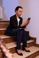 Stressed exhausted overworked businesswoman working on job deadling using smartphone overtime. Serious entrepreneur working on job project sitting on staircase of business building at night hours for job. photo