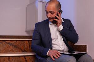 Exhausted overworked businessman talking on phone with team about project deadline.Entrepreneur working late in evening corporate sitting on stairs in building office. photo