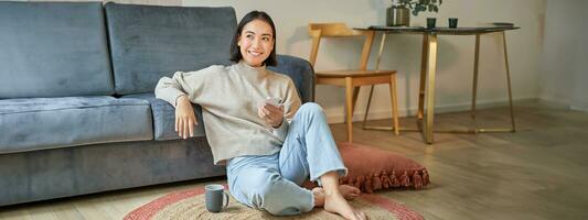 Portrait of stylish korean woman sits on floor with smartphone, using mobile phone, smiling pleased, concept of staying at home and relaxation photo