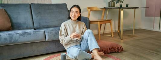 Image of stylish young woman in modern house, using mobile phone, sitting on floor and holding smartphone, drinking from cup photo