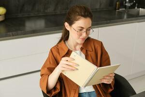 cerca arriba retrato de joven mujer, estudiante en cocina, participación computadora portátil, revisando para examen a hogar, estudiando, leyendo su planificador foto